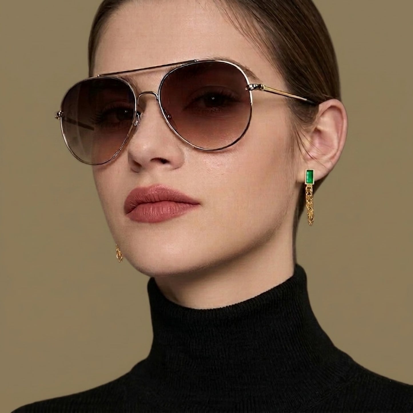 Woman wearing sunglasses and a black turtleneck wearing green golden chain earrings on a golden background